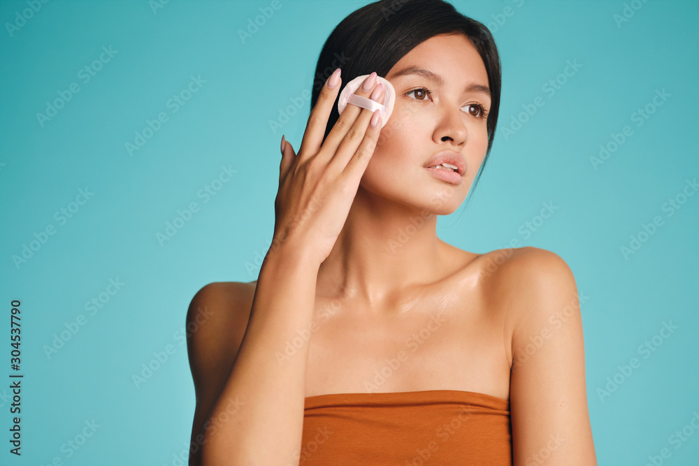 Attractive Asian brunette girl dreamily using powder puff posing over colorful background isolated Attractive Asian brunette girl dreamily using powder puff posing over colorful background isolated