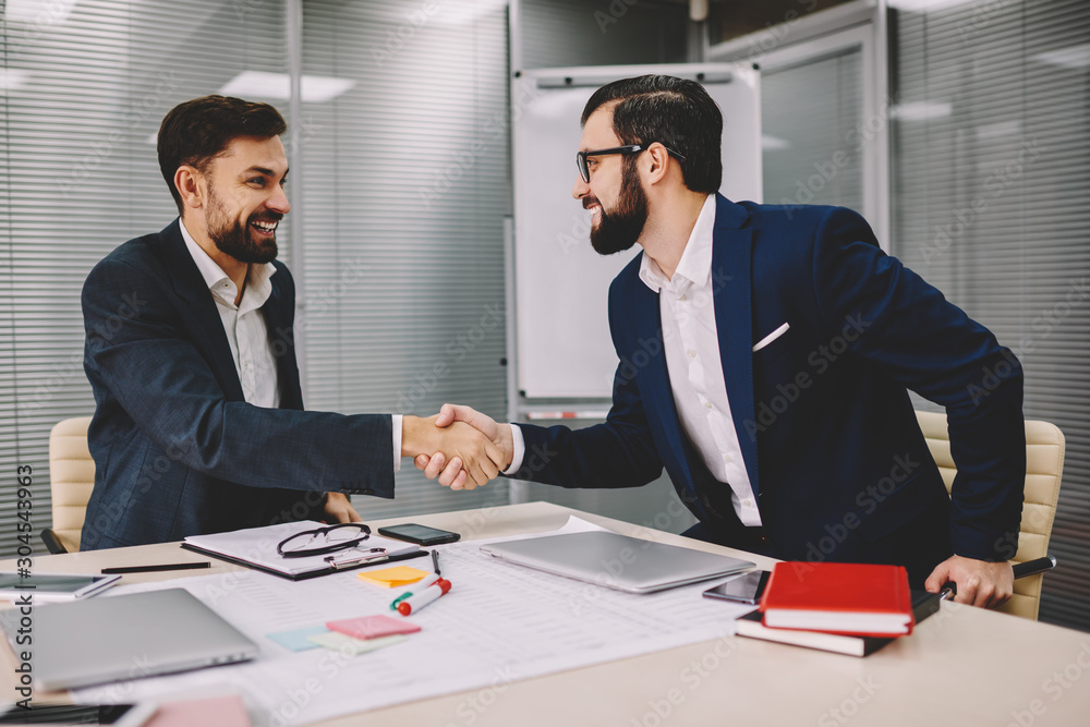 Two successful businessmen dressed in formal wear shaking hands gesturing prosperous partnership of finance company during meeting in office.Happy professional traders have deal about cooperation