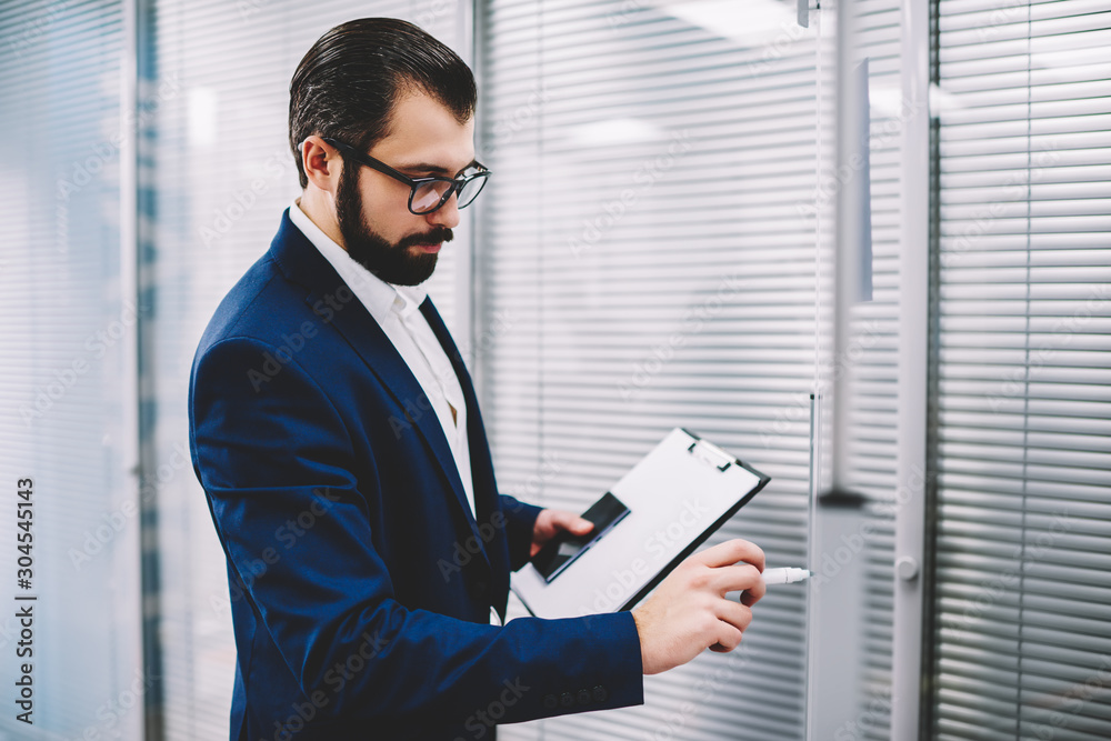 Fototapeta premium Pensive bearded proud ceo with marker in hands drawing schedule of financial reports.Concentrated executive director dressed in formal wear writing information of working process standing in office