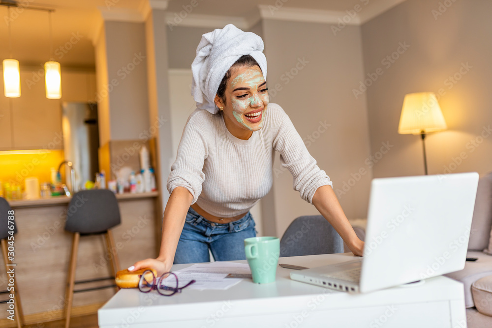Beautiful young woman working on laptop computer while sitting at the living room, drinking coffee with face mask. Beautiful young woman smiling with face mask and looking at laptop screen Beautiful young woman working on laptop computer while sitting at the living room, drinking coffee with face mask. Beautiful young woman smiling with face mask and looking at laptop screen