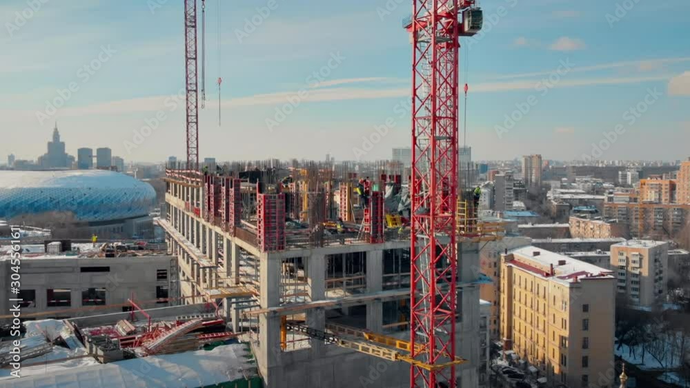 Picturesque aerial view to the construction works on the top of a skyscraper on a sunny winter day. Giant cranes, workers, empty window holes of a new building and urban panorama on the background. Picturesque aerial view to the construction works on the top of a skyscraper on a sunny winter day. Giant cranes, workers, empty window holes of a new building and urban panorama on the background.