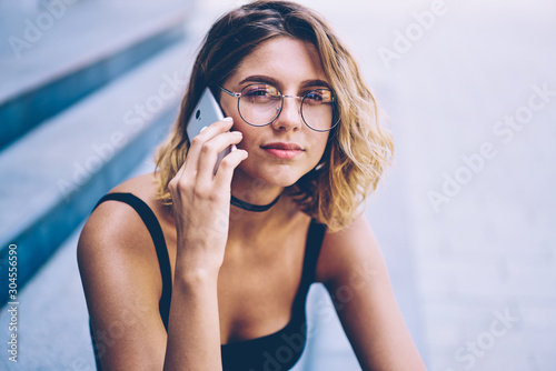 Close up portrait of attractive hipster girl in trendy sunglasses looking at camera during international smartphone conversation, charming woman calling to operator during free time outdoors
