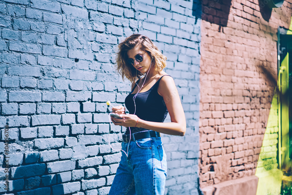 Trendy female meloman searching new audio tracks via media application on cellular for listening music on leisure, young hipster girl in electronic headphones enjoying music playlist during free time