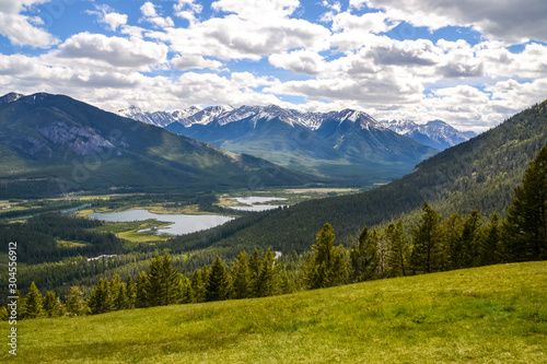 A view of the green grassy hills overlooking Banff mountain range with the valleys below covered trees and wetlands on a partially sunny day in Banff, Canada. © Hal Photography