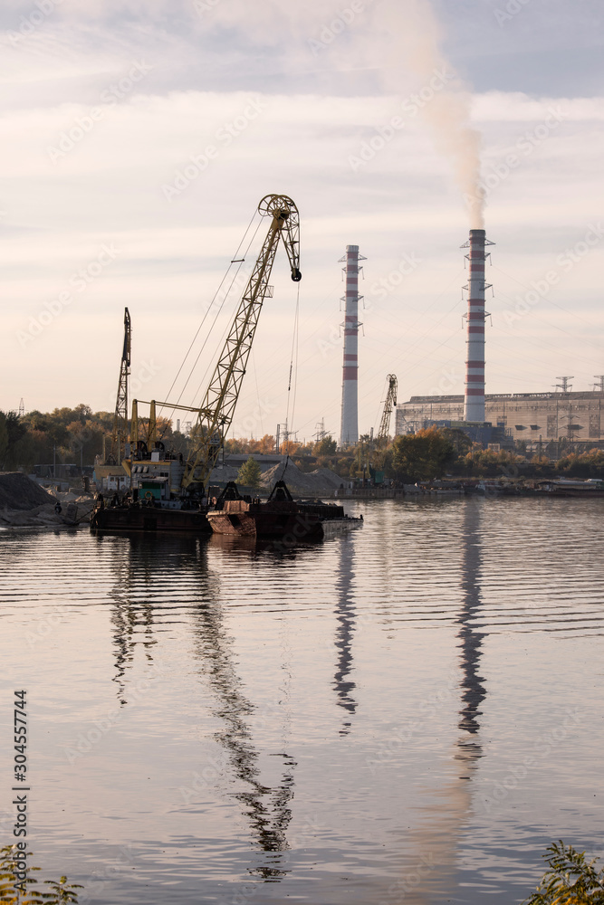 Fototapeta premium Industrial landscape with cranes and pipes. The plant works near the river. Crane on a barge loads cargo. Industrial area. Cityscape Scenery.