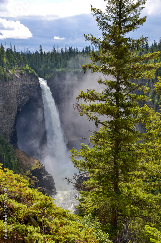 Helmcken Waterfall towers 141 meters above Murtle River with a constant flow of water gushing over the rocky cliff face feeding the dense pine forest it sits within on a partially sunny day in Canada.