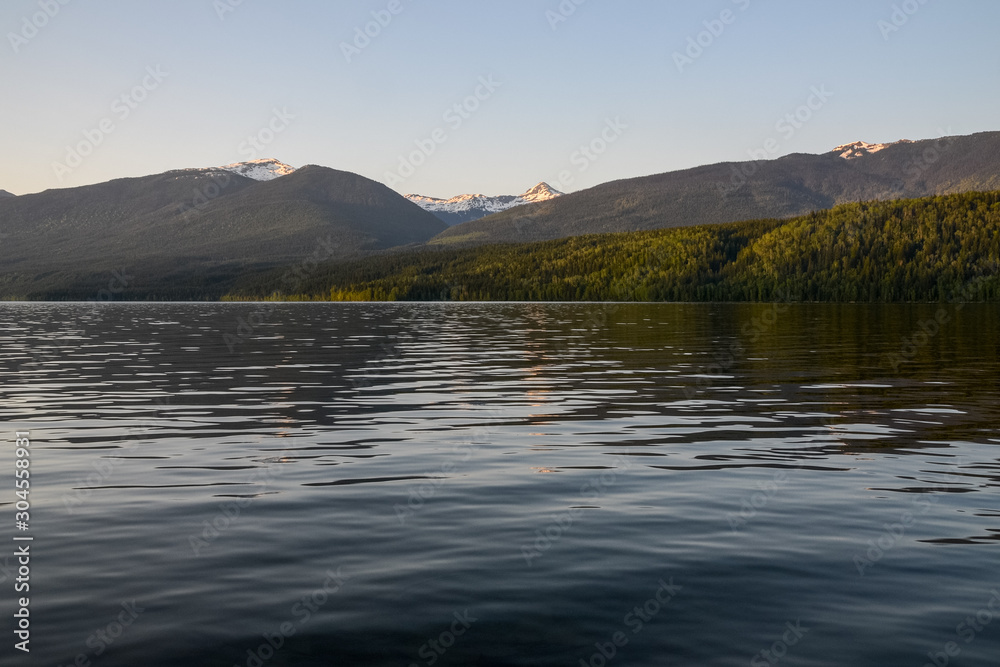 A warm sunset glow tints the clear sky, highlighting the snow capped mountain peaks and dense pine forest beyond the reflective lake, skewed its rippled surface.
