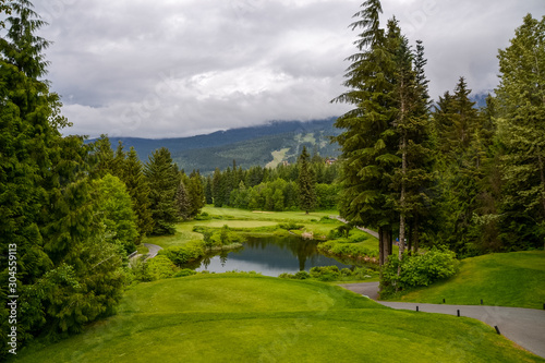 A gravel path wraps around the teeing greens with perfectly landscaped vibrant green grass and a small reflective pond. Low cloud hovers over the surrounding pine forest on this grey day.