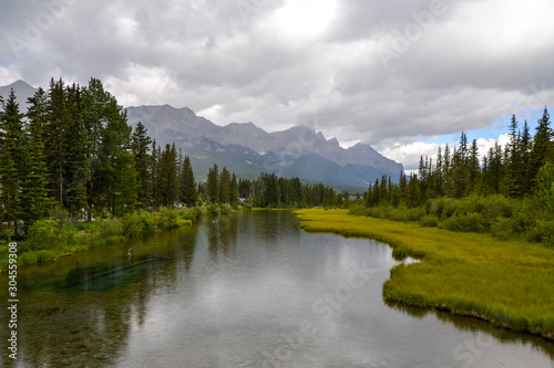 The clear water of Bow River with light droplets of rain on the surface. The clouds skim across the top of the rocky Mountain range in the distance on this cold and wet day in Canada.