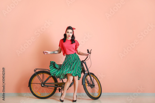 Fototapeta Naklejka Na Ścianę i Meble -  Portrait of beautiful tattooed pin-up woman with bicycle near color wall