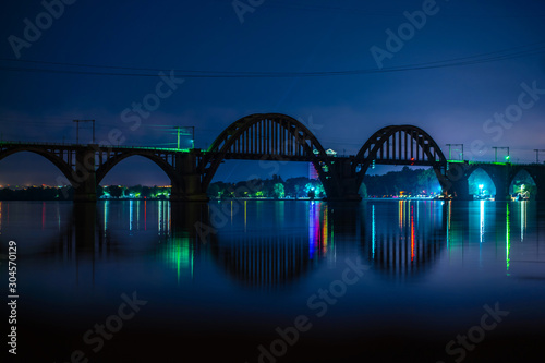 night landscape of the city promenade with many colored lights reflected in the water. Silhouetted humpback railway bridge