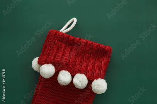 Red knitted Christmas stocking with white pom poms on green background, copy space