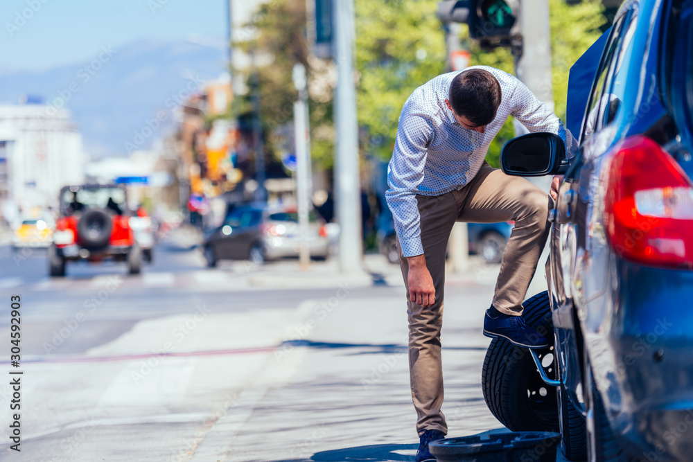 A strong businessman is bending his knee while trying to change a flattie on his car with his lug wrench. A strong businessman is bending his knee while trying to change a flattie on his car with his lug wrench.