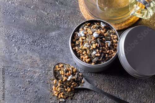 Organic and natural brown rice tea, genmaicha in a container. Accompanied by a cup of hot tea on gray background.