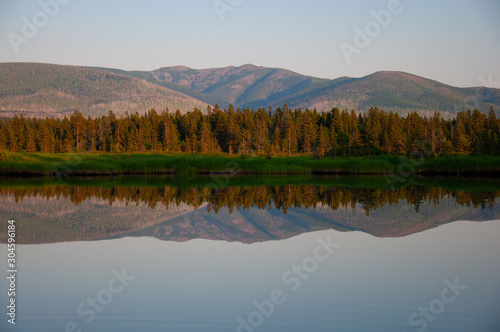 Evening photo of the lake. In the background is the taiga and the mountains. A beautiful reflection in the water.