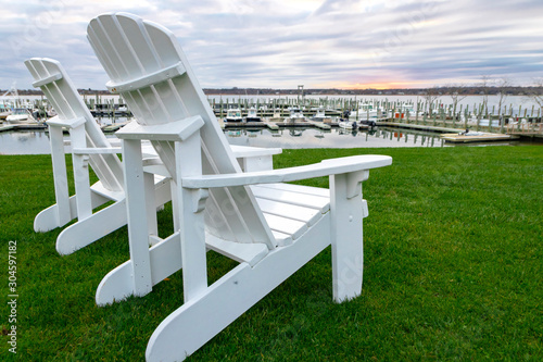 White "Adirondack" style lounge chairs on green grass at waterfront marina, with boats docked in background