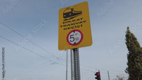 Yellow traffic sign in Turkish language. Sign indicates that there is electrical cables above. Blue sky, traffic lamp and a tree in background. photo