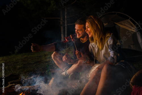Young couple baking sausages on the campfire and drinking beer in the forest hill in the dusk photo