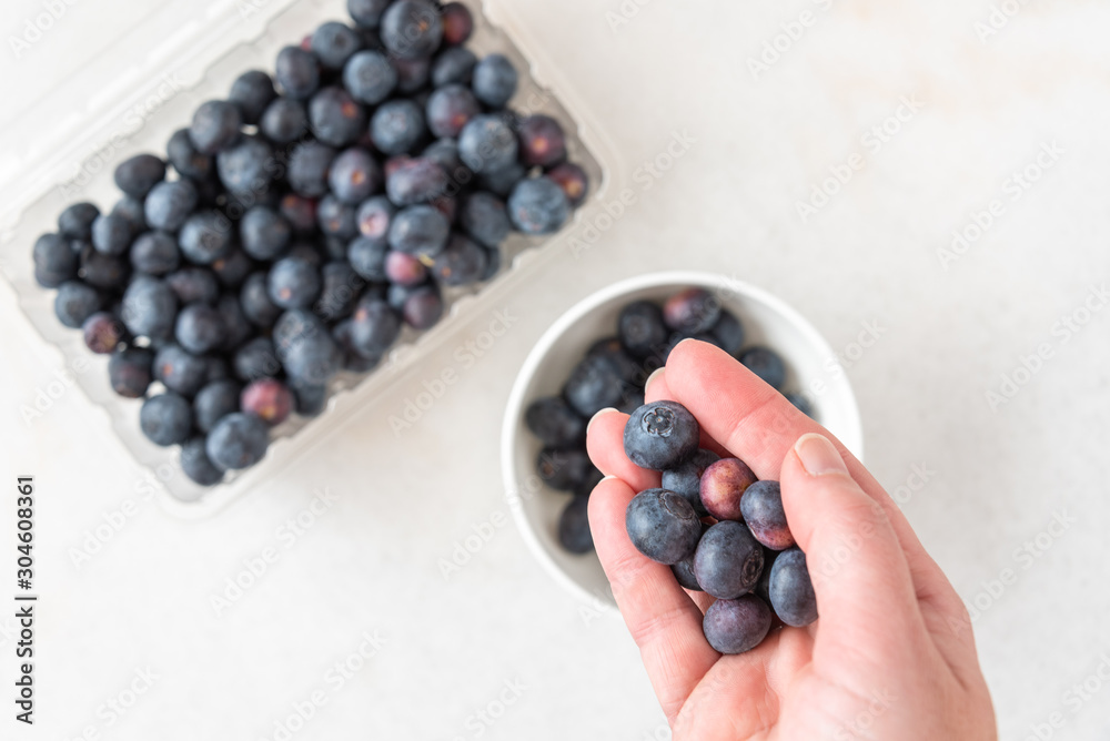 Woman’s hand picking blueberries from clamshell container on a white granite counter, small white bowl Woman’s hand picking blueberries from clamshell container on a white granite counter, small white bowl