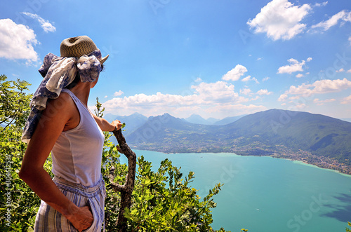 A young happy woman wearing a hat admires a panoramic view on the Lake Annecy from mont Veyrier to mont Baron hiking track, France