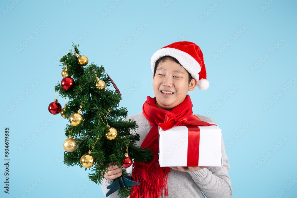 Happy Asian preteen boy holding Christmas tree and gift box on blue background, Christmas concept. Happy Asian preteen boy holding Christmas tree and gift box on blue background, Christmas concept.