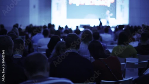 Unrecognizable businessman on stage talking microphone and showing presentation on screen to large audience sitting back to camera, blur background. Training, conference, public speech concept. photo