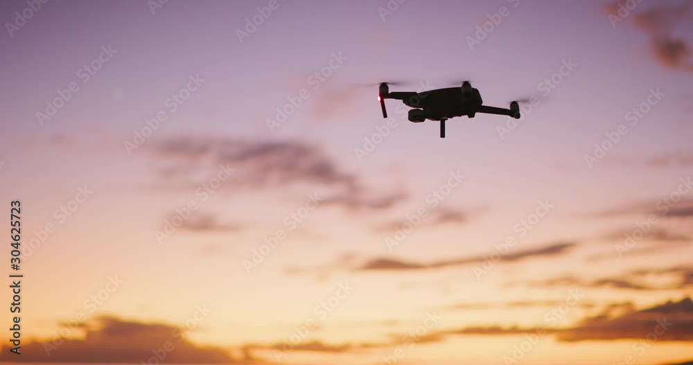 Orbiting view of a quadcopter drone silhouette hovering and flying at sunset with a purple yellow sky Orbiting view of a quadcopter drone silhouette hovering and flying at sunset with a purple yellow sky