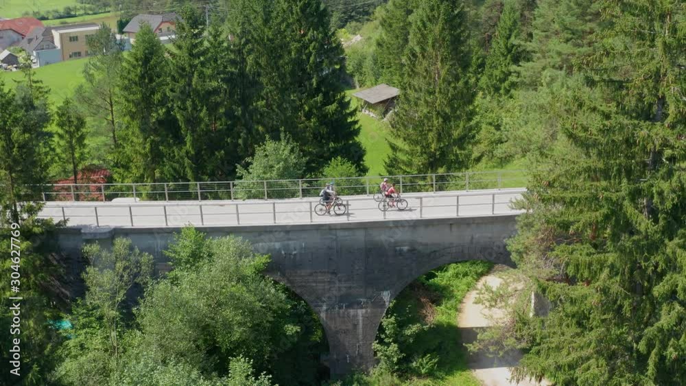 Aerial side shot of Caucasian family parents and children in bicycles crossing a bridge together in Slovenia, Drone orbits right as it tracks their movement Aerial side shot of Caucasian family parents and children in bicycles crossing a bridge together in Slovenia, Drone orbits right as it tracks their movement