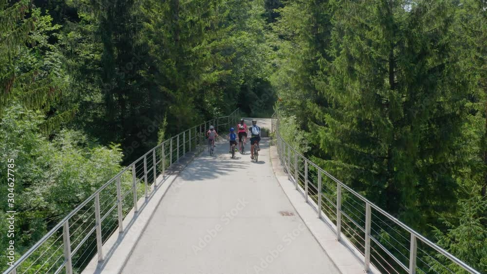 Aerial shot of Caucasian family parents and children in bicycles crossing a bridge together in Slovenia, Drone lifts at the end to reveal the forest ahead Aerial shot of Caucasian family parents and children in bicycles crossing a bridge together in Slovenia, Drone lifts at the end to reveal the forest ahead