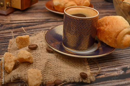 A Cup of coffee  pieces of brown sugar and a croissant on a wooden background.