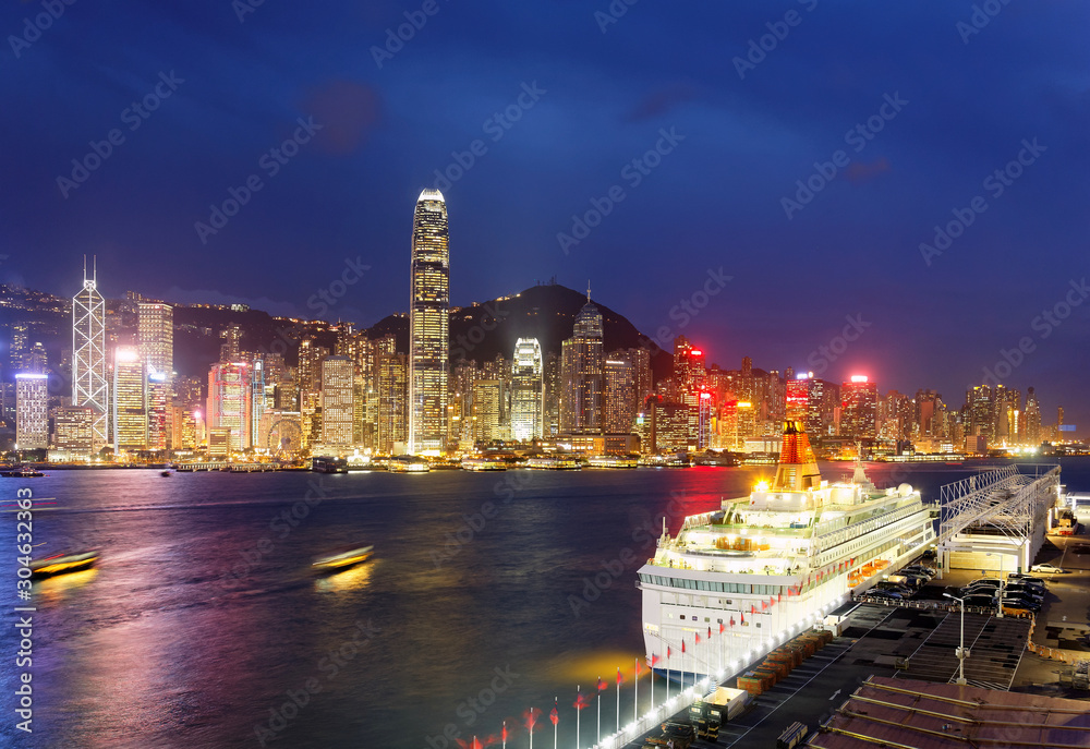 Night skyline of Hong Kong, with landmark IFC Tower standing amid skyscrapers by Victoria Harbour, a luxury cruise liner parking at Ocean Terminal in Tsim Sha Tsui and city lights glowing in blue dusk