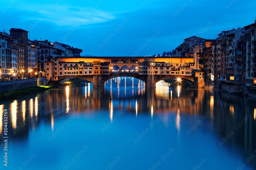 Morning scenery of historic Ponte Vecchio ( Old Bridge ), a beautiful landmark bridge over Arno River in Florence, Italy, with lights reflected in the peaceful water in the blue twilight
