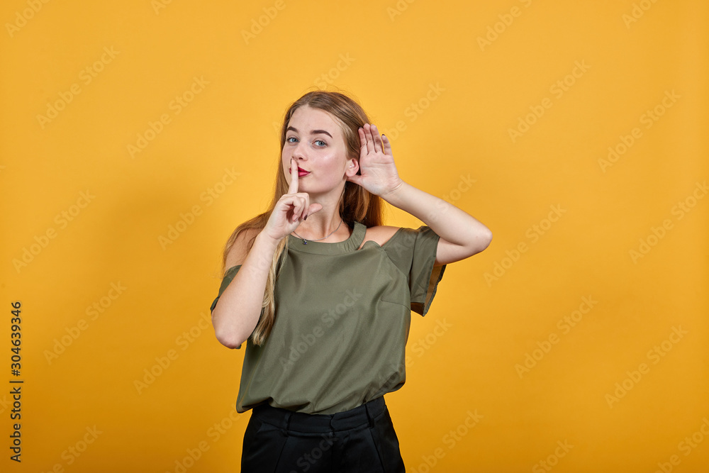 Portrait of cute smiling young woman in casual shirt putting hand prop up on ear, keeping finger on mouth, shh gesture isolated on orange wall background in studio. Portrait of cute smiling young woman in casual shirt putting hand prop up on ear, keeping finger on mouth, shh gesture isolated on orange wall background in studio.