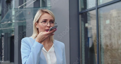 Caucasian young beautiful busy businesswoman walking outdoors in the morning and recording a voice message while talking to the phone speaker. Close up. photo