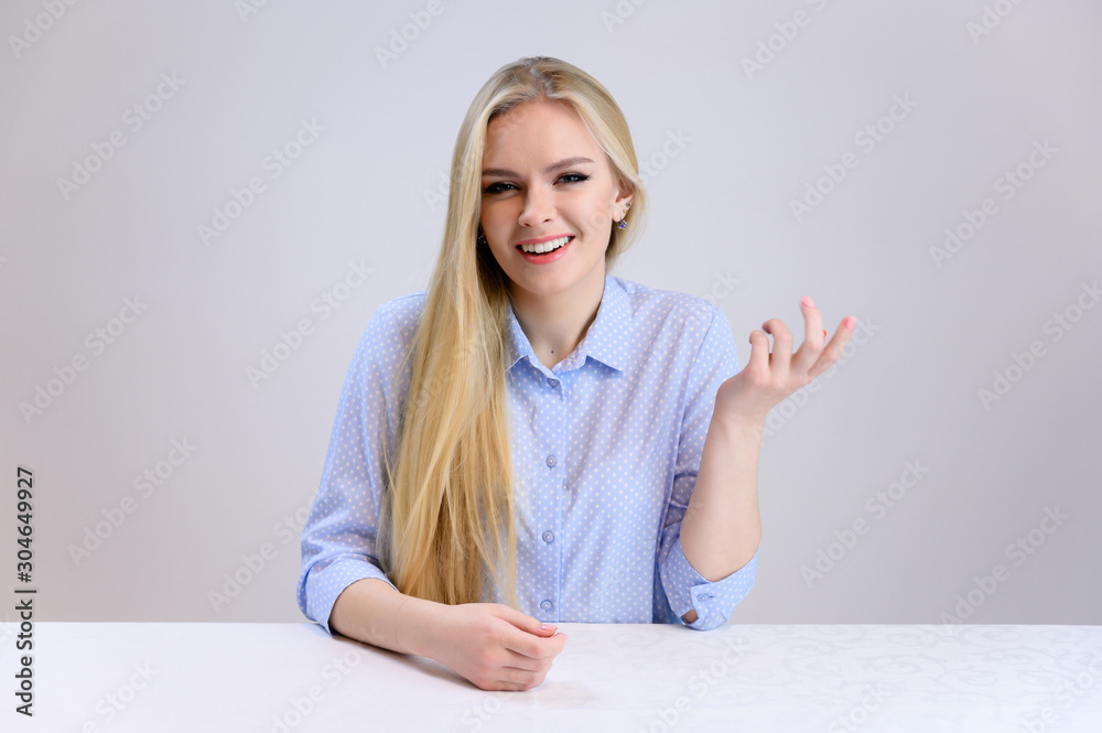 Concept cute model talking to the camera sitting at the table. Close-up portrait of a beautiful blonde girl with excellent makeup with long smooth hair on a white background in a blue shirt. Concept cute model talking to the camera sitting at the table. Close-up portrait of a beautiful blonde girl with excellent makeup with long smooth hair on a white background in a blue shirt.