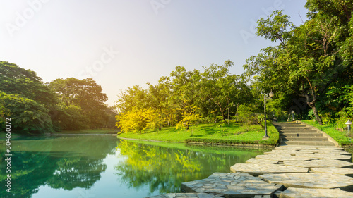 A lake in public park  greenery trees  shrub and bush  green grass lawn in a good care maintenance landscapes  decorated with arch curve concrete bridge cross the water under white clouds blue sky