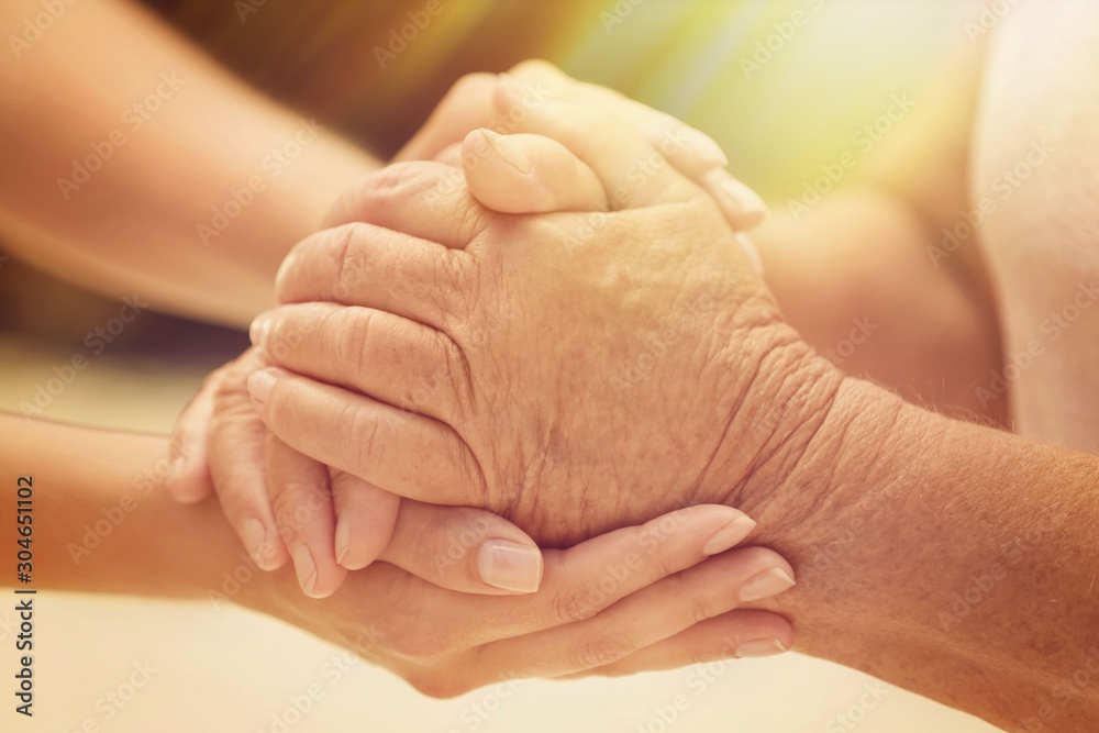 Fototapeta premium Volunteer and elderly woman holding hands in sunlit room, closeup