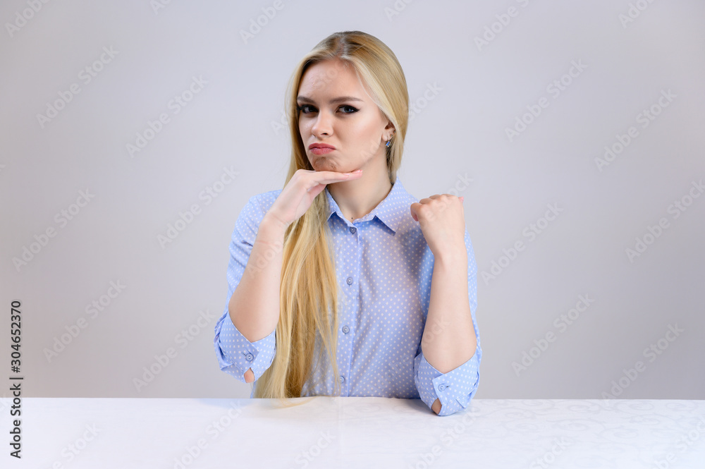 Concept cute model talking to the camera sitting at the table. Close-up portrait of a beautiful blonde girl with excellent makeup with long smooth hair on a white background in a blue shirt. Concept cute model talking to the camera sitting at the table. Close-up portrait of a beautiful blonde girl with excellent makeup with long smooth hair on a white background in a blue shirt.