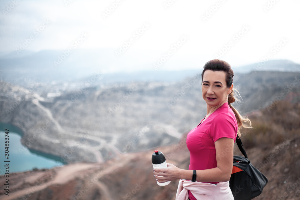 Miiddle aged woman drink water from sport bottle  outdoor over mountain  peaks and the lake. The traveller dressed sport wear.