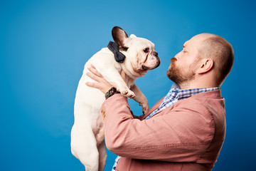 Bald man in jacket with french bulldog in his arms on empty blue background