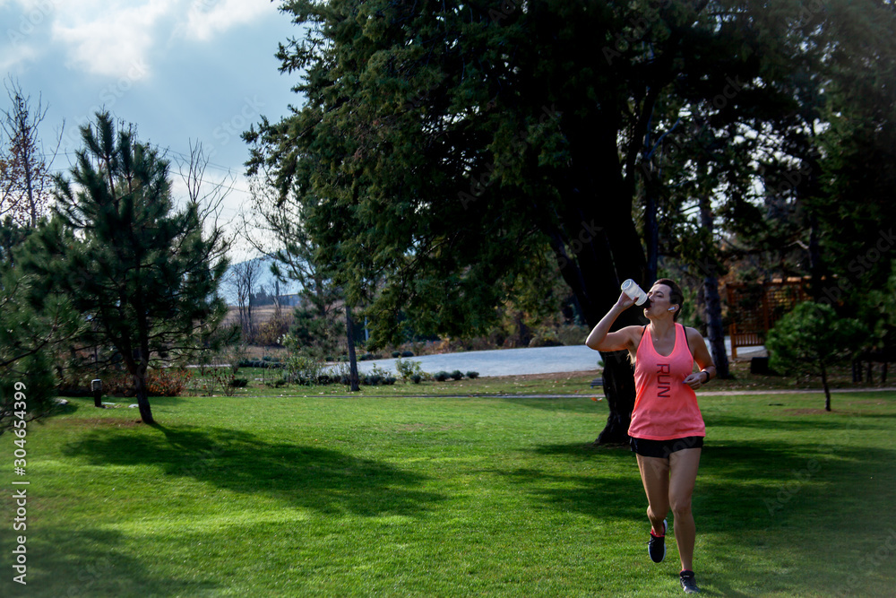 40s years old active woman dressed sportwear and headphones is running and drink water outdoor in the park with green grass. Mature woman living healthy lifestyle 40s years old active woman dressed sportwear and headphones is running and drink water outdoor in the park with green grass. Mature woman living healthy lifestyle