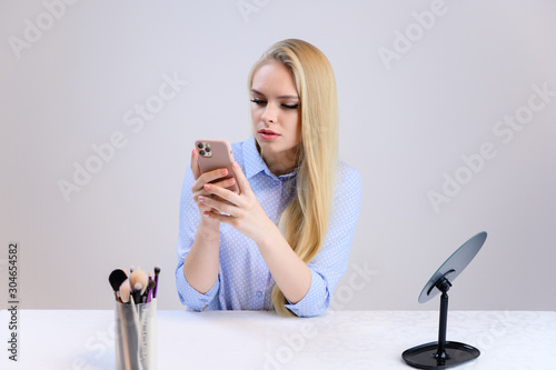 Concept cute model puts makeup on herself sitting at the table. Close-up portrait of a beautiful blonde girl with excellent makeup with long smooth hair on a white background in a blue shirt.