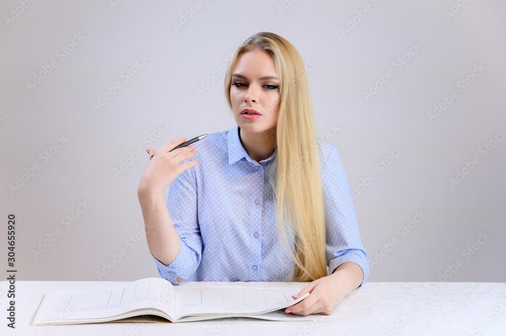 Concept cute model student secretary works sitting at a table. Close-up portrait of a beautiful blonde girl with excellent makeup with long smooth hair on a white background in a blue shirt. Concept cute model student secretary works sitting at a table. Close-up portrait of a beautiful blonde girl with excellent makeup with long smooth hair on a white background in a blue shirt.