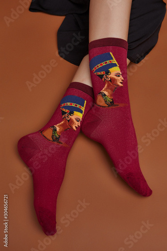Cropped shot of a girl's foots, lying on a brown background. It is burgundy Nefertiti print socks with dark stripes on a sock's top on foots. 