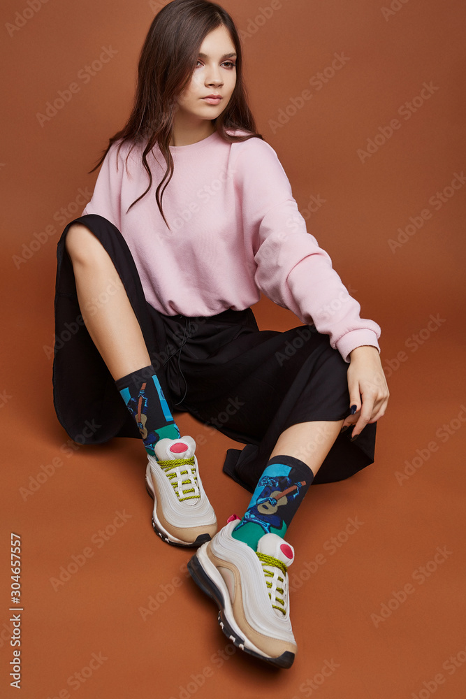 Full length shot of a dark-haired girl, sitting on a floor. She wearing  pink sweatshirt, black culottes, emerald artwork socks with man playing  guitar print and white sneakers. Stock Photo | Adobe