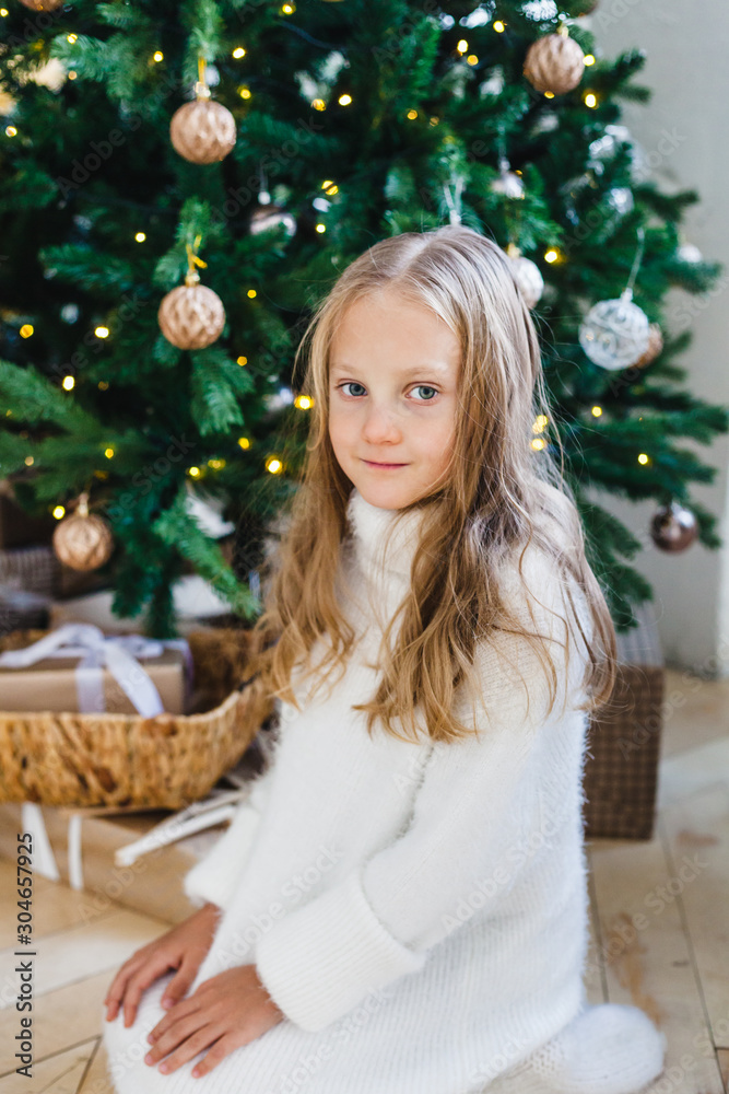 Girl near the Christmas tree decorated with balls and garland, girl with long hair in a white sweater, waiting for the new year and Christmas, gifts
