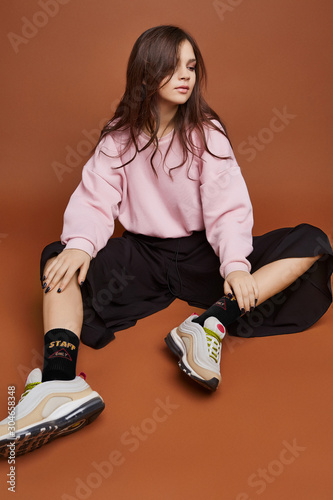 Full length shot of a dark-haired girl, sitting on a floor. She wearing pink sweatshirt, black culottes, black artwork socks with lettering 