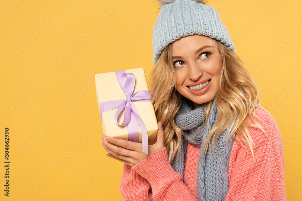 Smiling woman in hat and scarf holding gift box isolated on yellow Smiling woman in hat and scarf holding gift box isolated on yellow
