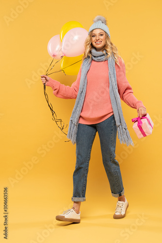 Smiling woman in hat and scarf holding balloons and gift box on yellow background © LIGHTFIELD STUDIOS