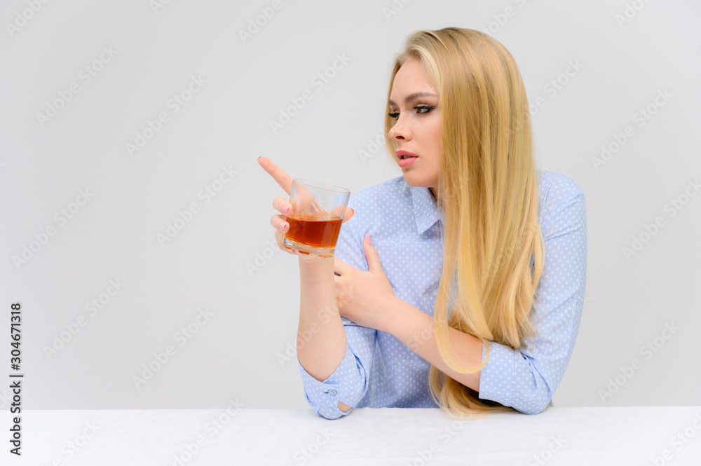 Concept woman with alcohol problems sits at a table with whiskey in a glass. Portrait of a beautiful blonde girl with excellent makeup with long smooth hair on a white background in a blue shirt. Concept woman with alcohol problems sits at a table with whiskey in a glass. Portrait of a beautiful blonde girl with excellent makeup with long smooth hair on a white background in a blue shirt.